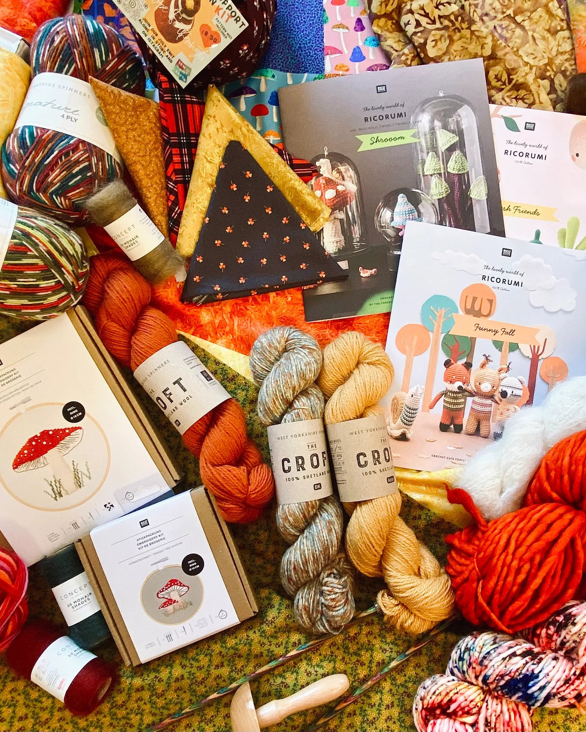 A flat-lay collection of knitting and crochet supplies, including multiple skeins of yarn in various colors, a crochet hook, pattern books, and a cozy knit scarf, all arranged on a textured green and yellow patterned fabric background.